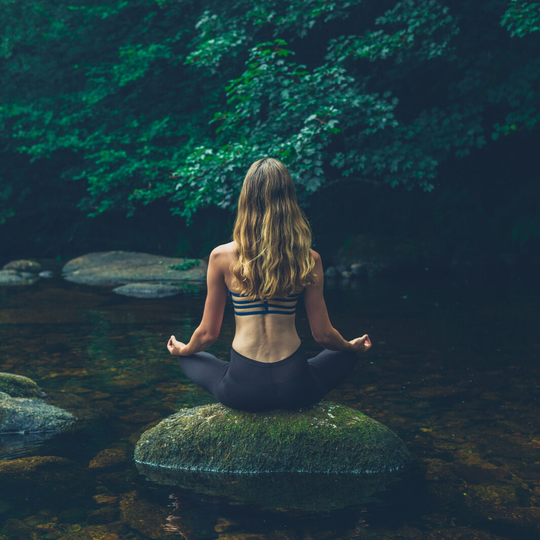 A young woman is meditating on a rock in the river